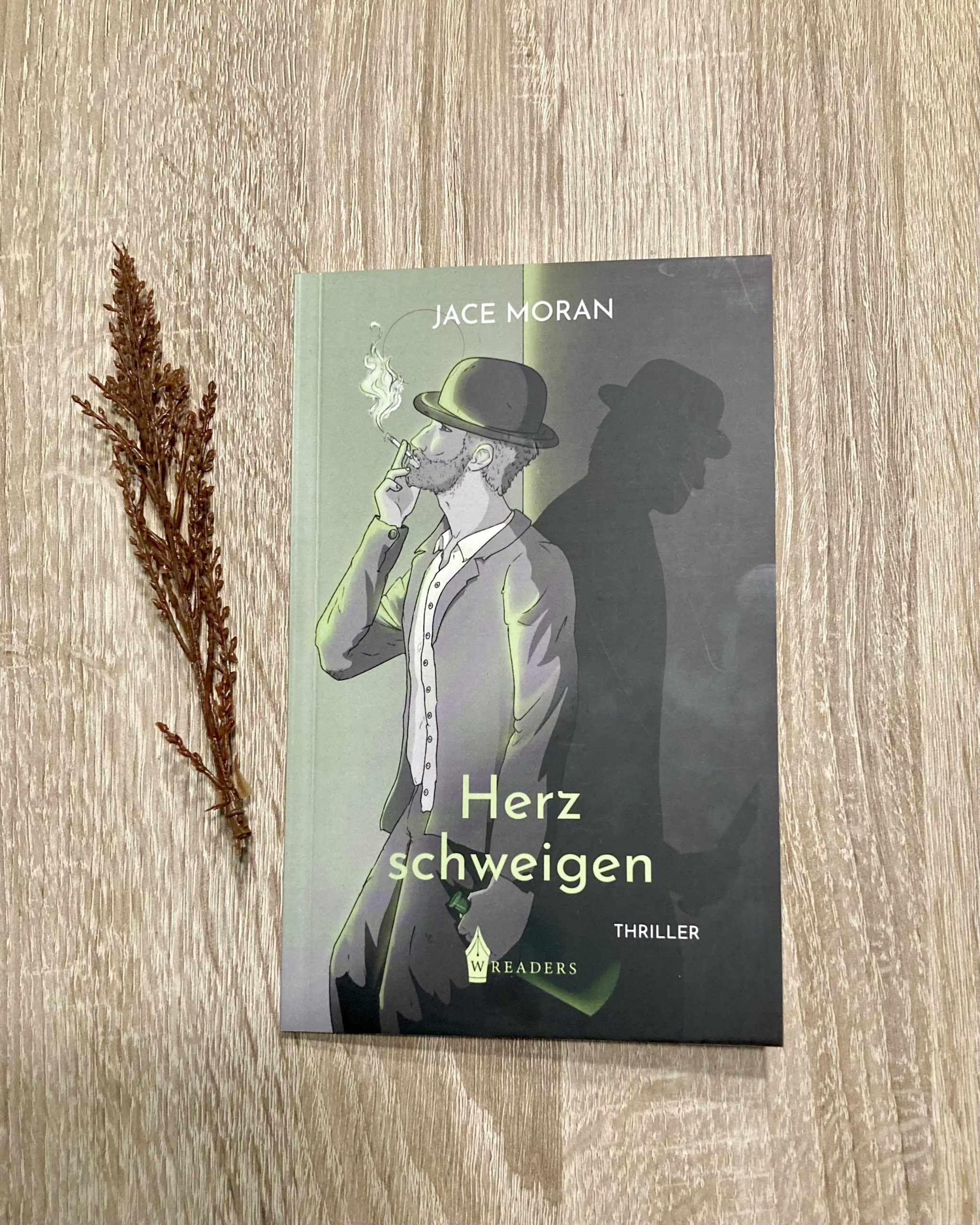 Herzschweigen von Jace Moran. Auf dem Cover ist die Zeichnung eines Manes mit Anzug und Hut zu sehen, der gegen eine Wand gelehnt eine Zigarette raucht. Von vorn wird er von grünem Licht beschienen; hinter ihm steht sein Schatten, der ein Messer in der Hand hält. Unten ist das Logo des W-Readers Verlag abgebildet.