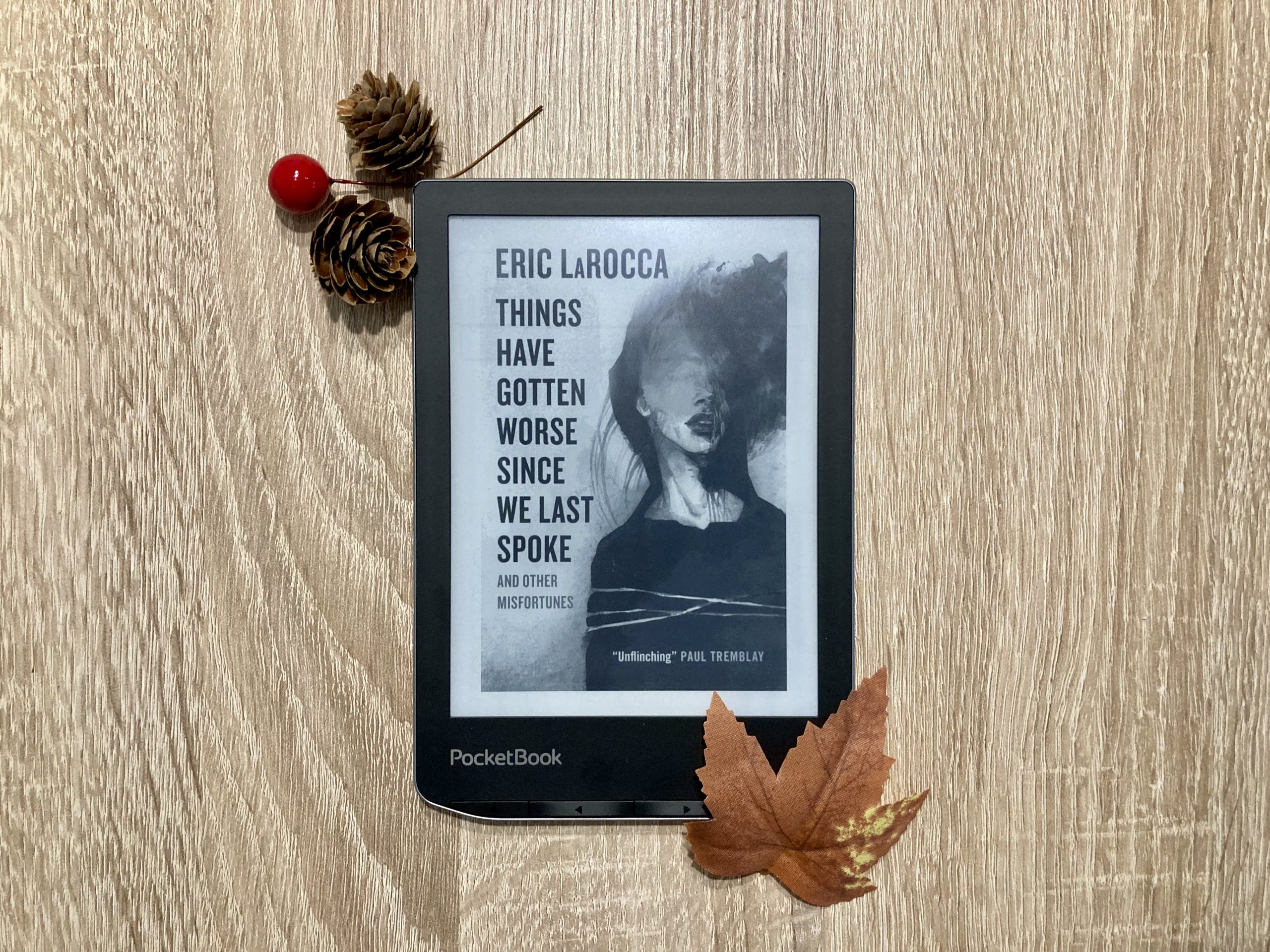 "Things Have Gotten Worse Since We Last Spoke And Other Misfortunes" von Erica LaRocca auf einem PocketBook E-Reader. dekoriert mit Tannenzapfen und einem Herbstblatt. Titel und Autor*in stehen untereinander in großen Buchstaben auf der linken Seite. Rechts ist die Zeichnung einer Frau abgebildet, deren Gesicht von den Augen aufwärts verwischt ist. Unten rechts steht der Kommentar "Unflinching" von Paul Tremblay.