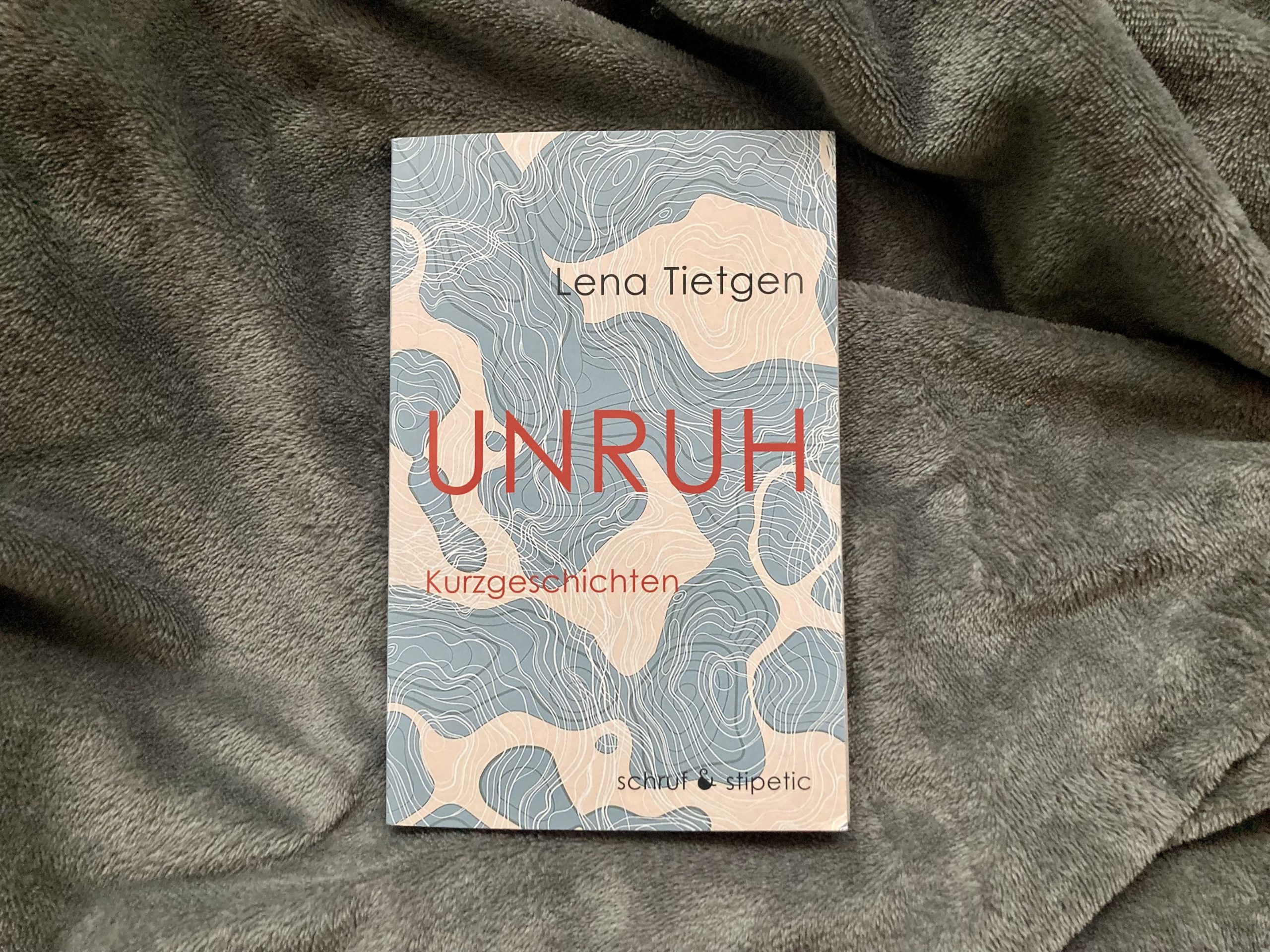 Das Buch "Unruh" von Lena Tietgen vor einem grauen Hintergrund. Das Cover besteht aus abstrakten blauen Formen, die vor einem beigefarbenen Hintergrund ineinander laufen. In der Mitte steht in großen roten Buchstaben der Titel "Unruh", darunter in kleinen roten Buchstaben der Untertitel "Kurzgeschichten". Über dem Titel steht in schwarzer Schrift der Name "Lena Tietgen", ganz unten rechts der Name des Verlags: Schruf und Stipetic.