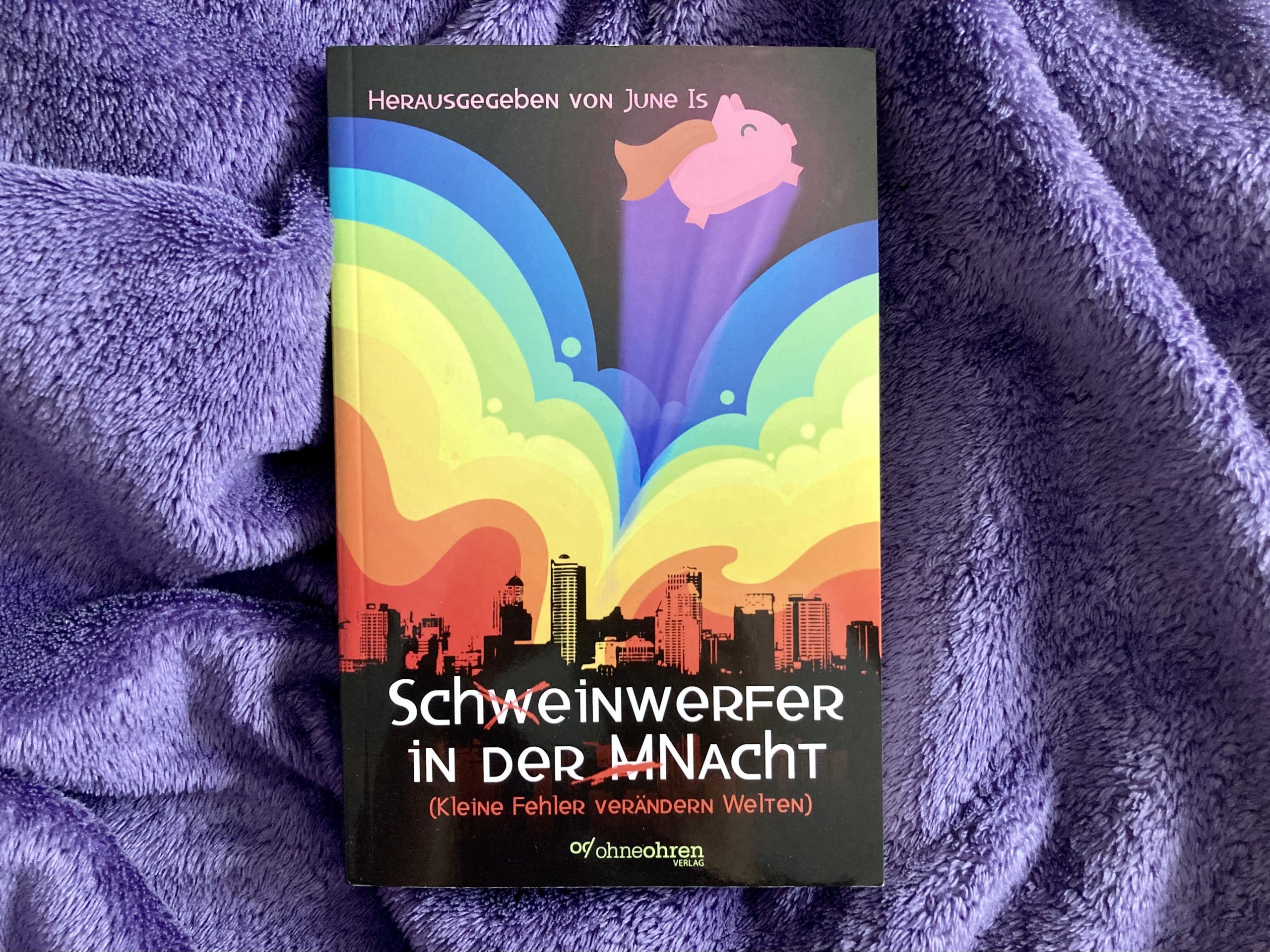 Ein Buch mit dem Titel „Schweinwerfer in der Macht“, wobei die Worte durch Durchstreichungen so korrigiert sind, dass er auch als „Scheinwerfer in der Nacht“ gelesen werden kann. Auf dem Cover ist die Silhouette einer Großstadt zu sehen, über der sich Wolken in Regenbogenfarben türmen. Aus diesen Wolken springt ein zufrieden schauendes Schwein mit Cape. Oben steht: „Herausgegeben von June Is“. Unten steht der Titel und der Untertitel „Kleine Fehler verändern Welten“. Darunter das Logo des Ohne-Ohren-Verlags.